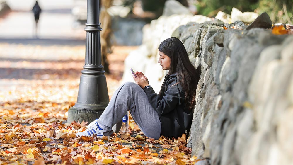 Faculty-&-Student-Picks-for-Fall--What-We’re-Watching-Now-_fall2025-home Ramapo College student sitting outside on the ground, surrounded by fallen autumn leaves, watching her phone