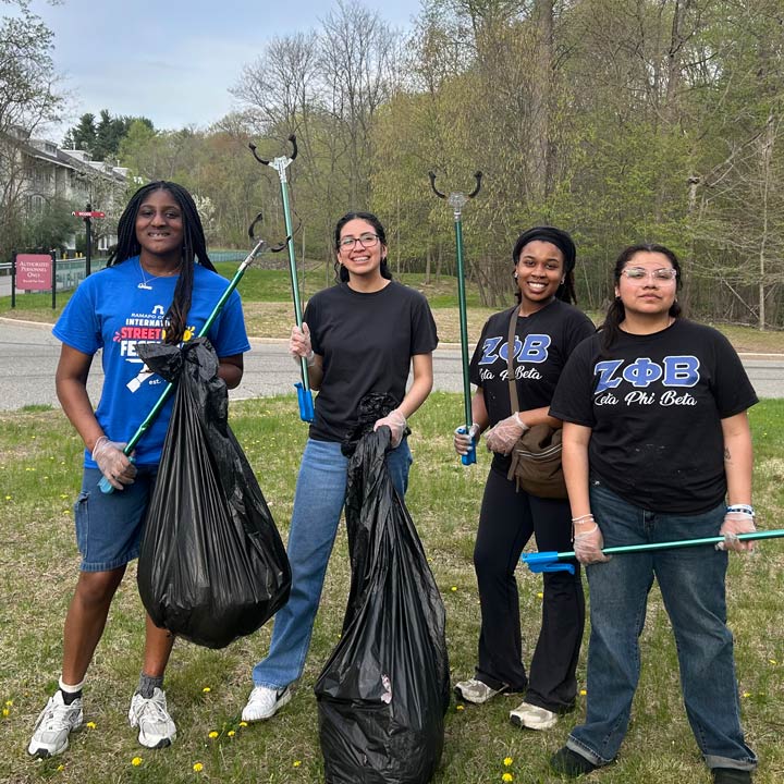 Four people stand outside holding trash bags and litter pickers, smiling after a cleanup event. Two wear matching Zeta Phi Beta shirts, and trees and a road are visible in the background.