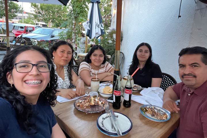 Edith Torres with her family are smiling and sitting around an outdoor restaurant table with food and drinks.