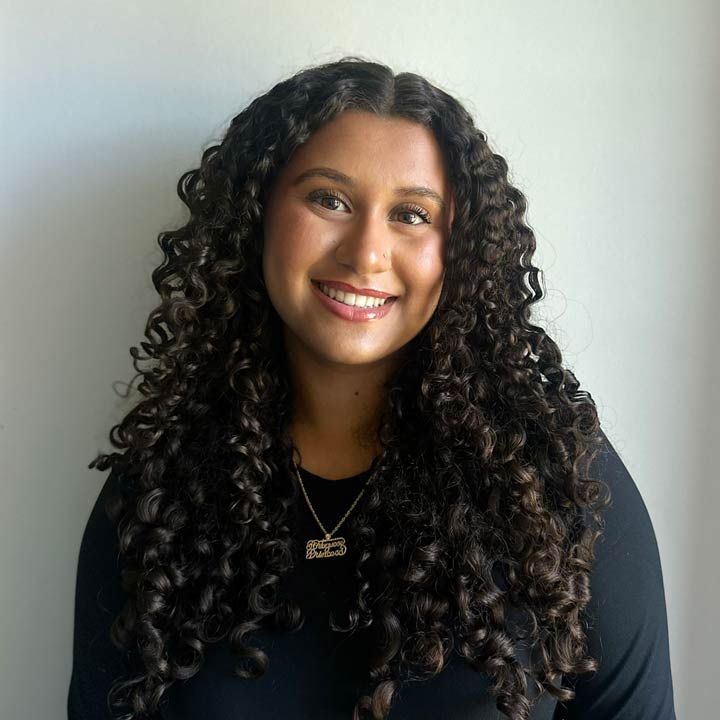 Kaylee Peña with long, dark, curly hair smiles at the camera. She is wearing a black top and a gold necklace, standing in front of a plain light-colored background.
