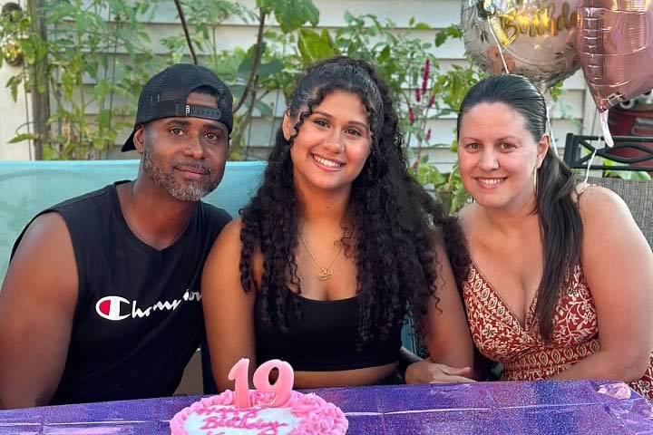 Kaylee Peña sitting Kaylee Peña with her parents, smiling at an outdoor birthday celebration.