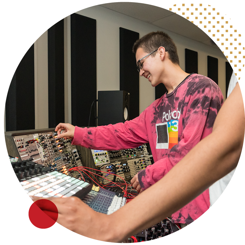 Two people, one in a pink tie-dye shirt, are using electronic audio equipment and synthesizers in a sound studio. One person is smiling and adjusting knobs, while the other’s arm is visible in the foreground.