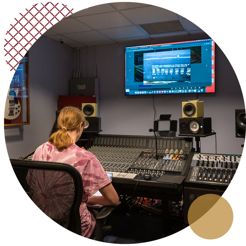 Person with light brown hair in a pink tie-dye shirt sits at a large audio mixing console in a recording studio, facing a monitor displaying sound editing software.