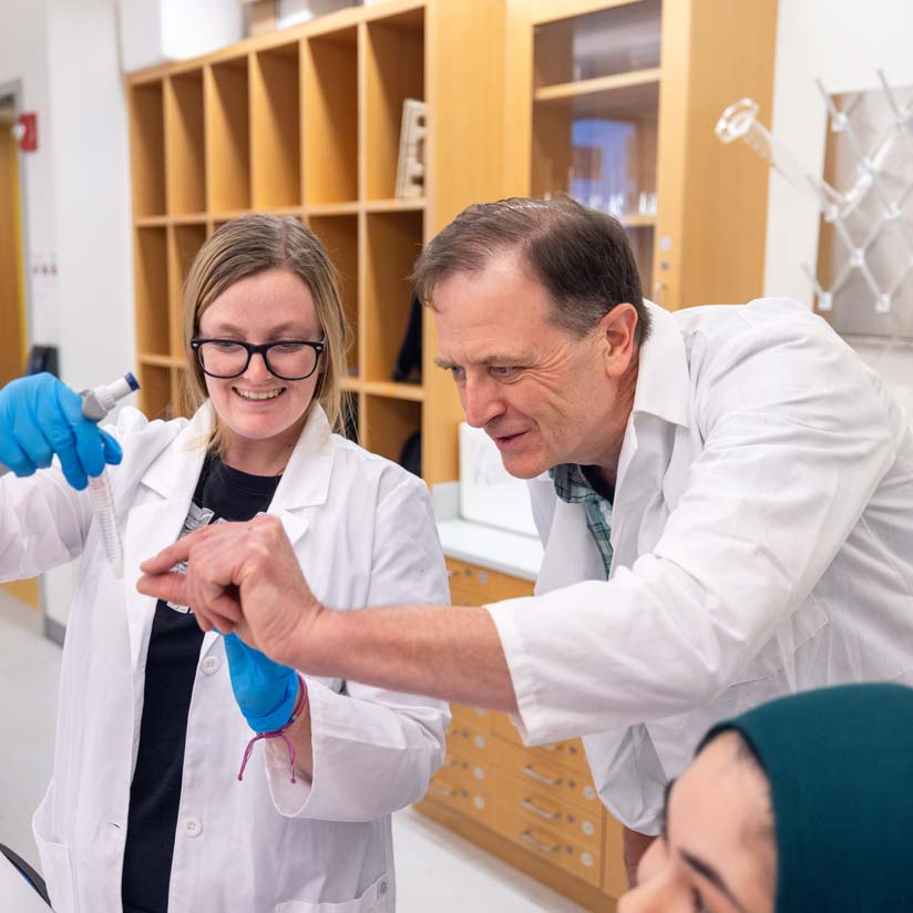 Professor Ashley Stuart in a lab coatsand gloves working with a Ramapo College student in a laboratory.