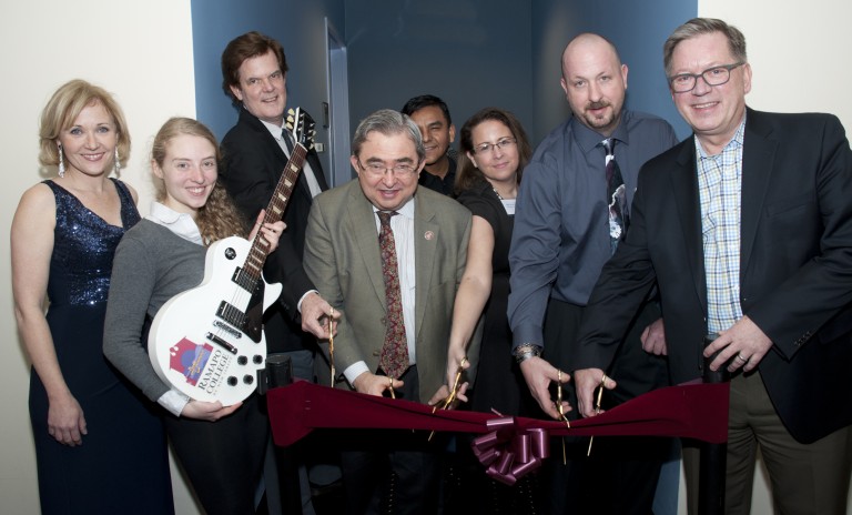 Eight people smile at a ribbon-cutting ceremony, with three men in front holding large scissors and one woman holding a white electric guitar. The group stands in a hallway with a maroon ribbon and bow.