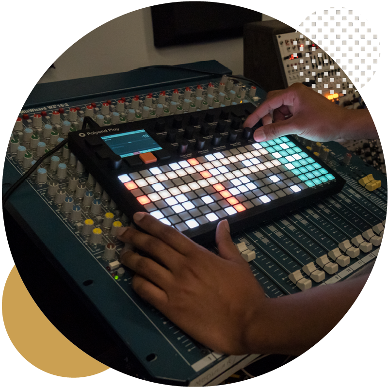 A person operates a music production controller with illuminated pads and knobs placed on top of a large audio mixing console.