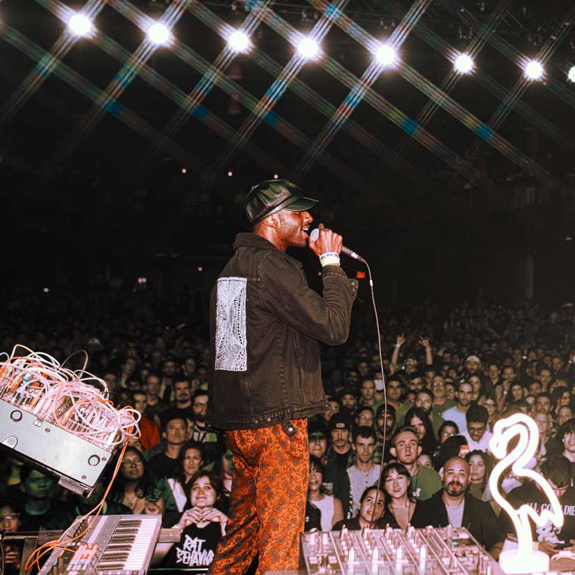 Ehiorobo Igiehon in a black jacket and cap sings into a microphone on stage before a large, energetic crowd, with bright stage lights overhead and electronic equipment and a flamingo light in the foreground.