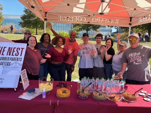 A group of parents and students stand under a canopy labeled Ramapo College, waving at the camera behind a table with snacks, bottled water, and a sign that reads The Nest. Its a sunny outdoor Homecoming event.