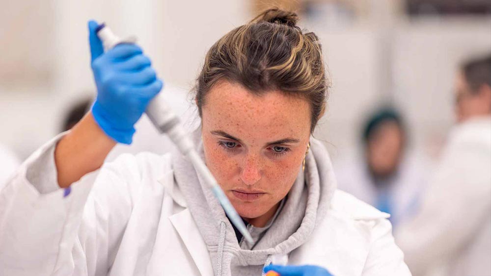 Ramapo College Bioinformatics student in a white lab coat and blue gloves uses a pipette to transfer liquid into a small container in a laboratory setting.