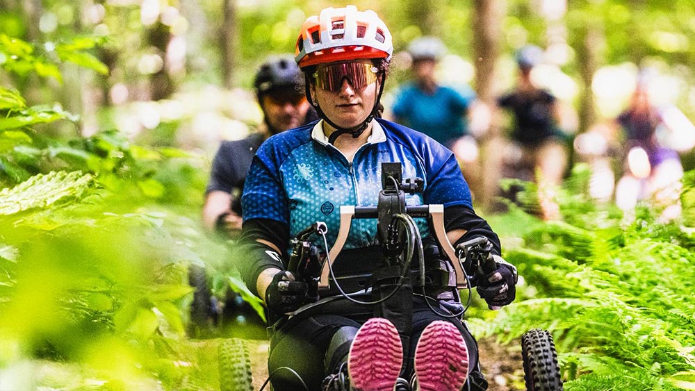 Emily Nappi’s ’25 wearing a helmet and sunglasses rides an adaptive mountain bike along a forest trail, surrounded by green foliage, with several other cyclists following behind.