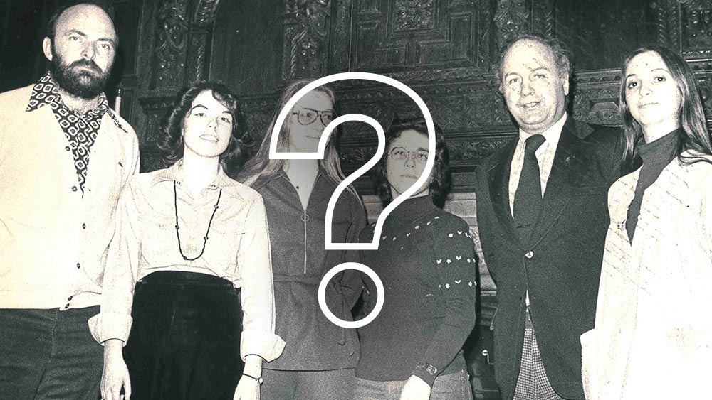 A black-and-white photo of Ramapo College constituents standing side by side in front of an ornate wooden background, all facing the camera and appearing to pose for a group portrait with a question mark infront