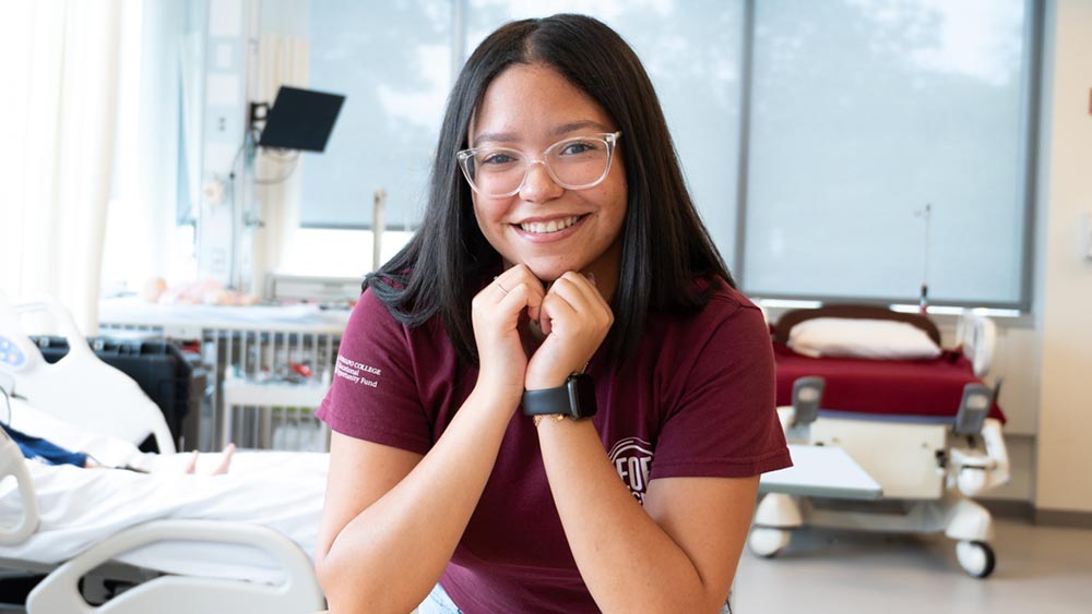 Ramapo College nursing student, Vanessa Garcia, with long dark hair and glasses smiles while sitting in a hospital room setting, wearing a maroon shirt and a smartwatch, with hospital beds and medical equipment in the background.