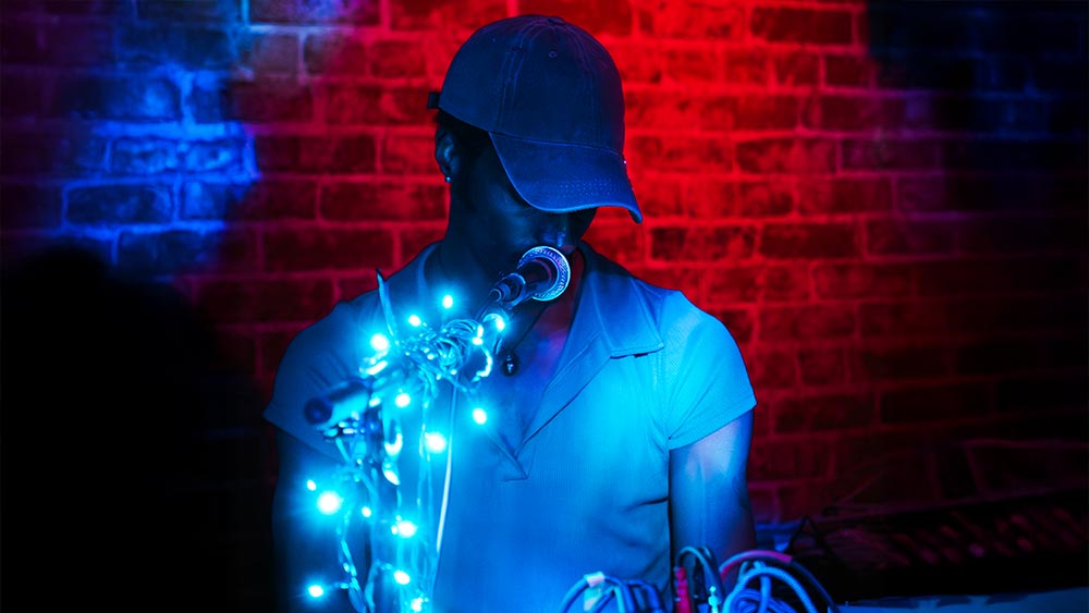 Ehiorobo Igiehon ’17, wearing a cap speaks into a microphone wrapped with blue lights, set against a red brick wall illuminated by red and blue lighting.