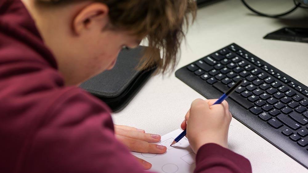 A person in a maroon sweatshirt draws shapes on white paper with a blue pen, sitting at a desk next to a black computer keyboard.