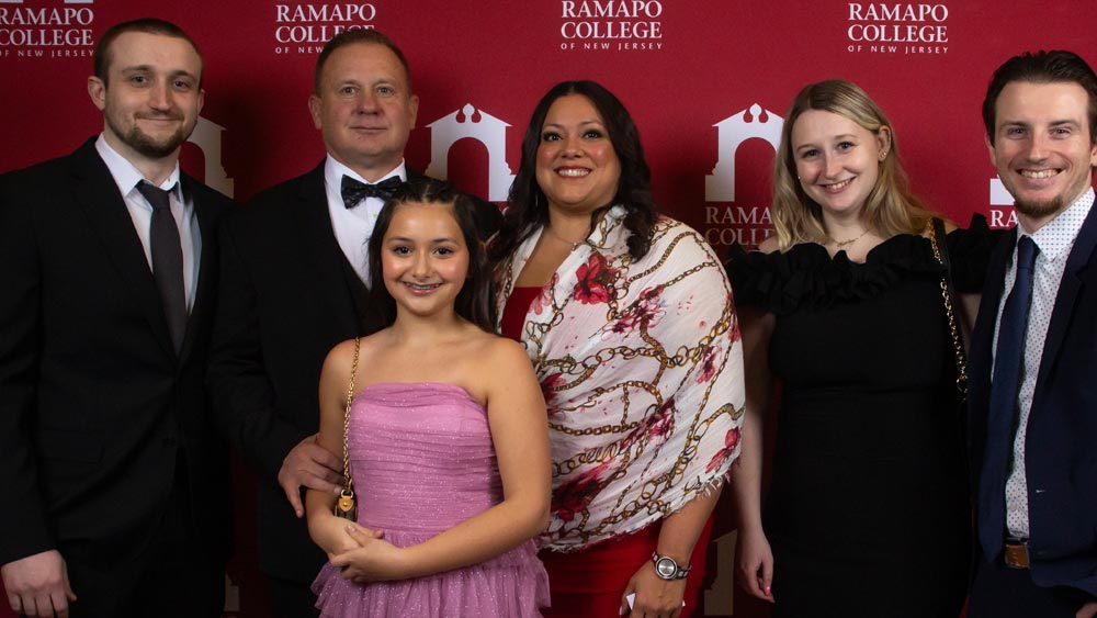 Six people dressed in formal attire stand smiling in front of a red backdrop with Ramapo College of New Jersey and its logo repeated across it.