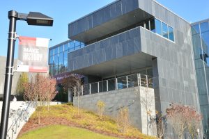 A side angle of the main entrance of the Learning Commons at Ramapo College. The core section of the building is slate and has a second story balcony. The rest is windowed exterior walls that reflect the sky and nearby campus spaces.