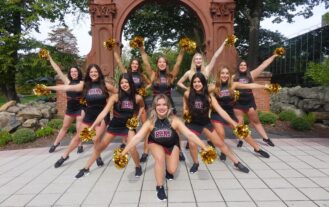 Ramapo College Dance Team in uniform with pom poms posing in front of The Arch.