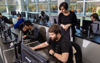 Prof. Nosrati stands behind a student working on a computer in the trading lab on campus.