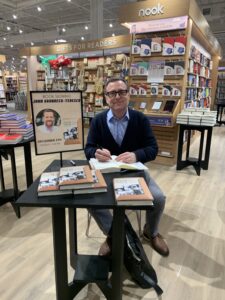 American Studies Professor John Gronbeck-Tedesco sits at a table signing a copy of his book inside Barnes & Noble bookstore.