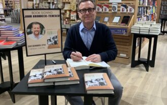 American Studies Professor John Gronbeck-Tedesco sits at a table signing a copy of his book inside Barnes & Noble bookstore.
