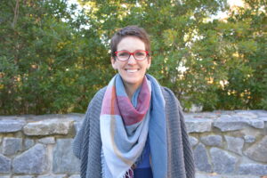 Susan Auger standing in front of trees wearing a colorful plaid scarf and red eyeglasses.