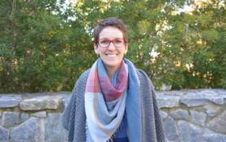 Susan Auger standing in front of trees wearing a colorful plaid scarf and red eyeglasses.