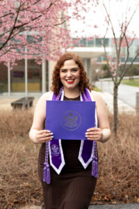 Anna Kozan smiles while wearing a purple nursing honor society graduation stole and holding her honor society certificate.