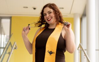Anna smiles while wearing a yellow nursing graduation stole and holding her stethoscope around her neck.