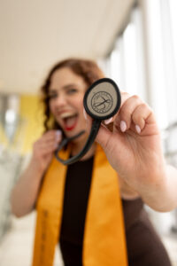 Anna playfully extends her stethoscope to the camera while laughing and wearing her yellow graduation stole.