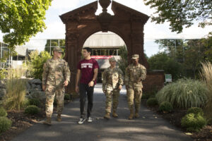 Four students walking toward the camera. Three are wearing green army fatigues and one is wearing a maroon Ramapo College VSO t-shirt. The Havemeyer Arch is in the background with green foliage.