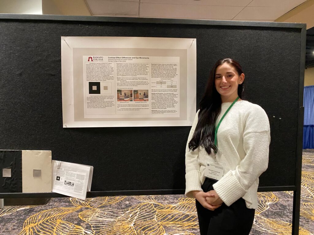 Alexandra Sacchinelli ‘24wears a white top and black pants and stands to the right of her poster featuring her research pinned to a black board.