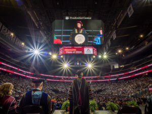 Image taken from the stage looking out over the arena floor filled with hundreds of graduates and seats filled with thousands of guests. The electronic banner on the big screen hanging from the ceiling reads "Congratulations Class of 2024!" President Jebb is standing at the podium. Her back is to the camera but she is facing the stage on the big screen.