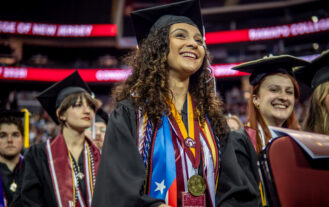 A smiling Stephanie Viera '25 is seated among graduates at the 2025 Commencement wearing a graduation robe, and several stoles, cords, and medals