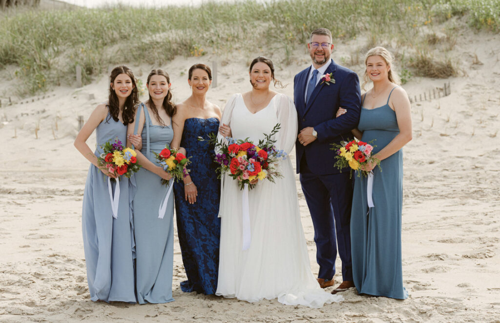 The Spizzo family stands on a beach for a family wedding photo with two daughters to the left of Tiffany and two daughters and her husband to the right. The four daughters all hold colorful floral bouquets.