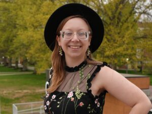A person in a black hat and black dress stands outside and smiles.