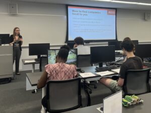 A career advisor presents job search advice in front of students. The advisor is at the front of a classroom and the students are facing the front of the classroom with their backs to us.
