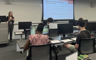 A career advisor presents job search advice in front of students. The advisor is at the front of a classroom and the students are facing the front of the classroom with their backs to us.