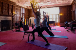Employees stand on yoga mats in a pose with arms lifted above their heads and legs in a split stance.