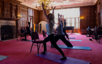 Employees stand on yoga mats in a pose with arms lifted above their heads and legs in a split stance.