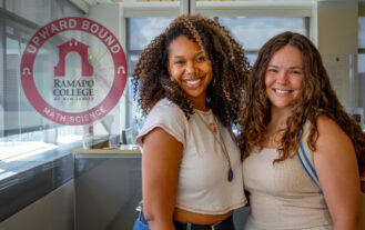 Two women who are graduate students stand next to each other in front of a sign for the Upward Bound office.