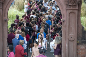 A single file line of college students file underneath an arch outside. The path leading up to the arch is lined with people congratulating them.