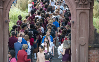 A single file line of college students file underneath an arch outside. The path leading up to the arch is lined with people congratulating them.