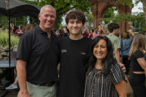 A  young man with curly brown hair is flanked by his parents. His father, in a black shirt stands on one side of him and his mother in a black and white blouse stands on the other.