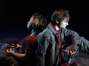 A female and male sit back to back on a stage. She is wearing a maroon sweater and he is wearing a jean jacket.