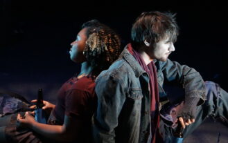 A female and male sit back to back on a stage. She is wearing a maroon sweater and he is wearing a jean jacket.