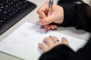 A hand holding a pen writes on a white piece of paper, while the other hand rests on the paper. A part of a keyboard is shown on the top left.