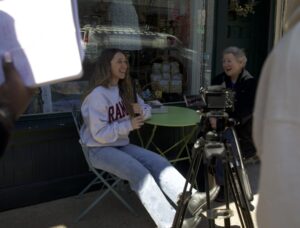 A female student wearing a Ramapo sweatshirt sits at a table outside a cafe with an older woman wearing a black coat. A camera is in front of them.