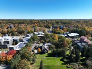 Aerial view of the Ramapo College campus. The sky is various shades of vibrant blue with trees bearing green needles and yellow, red, and orange leaves. A green grassy hill is in the foreground with the historic Birch Mansion to the right and the Academic Complex, topped by solar panels, to the left and center back.