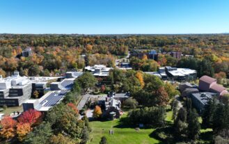 Aerial view of the Ramapo College campus. The sky is various shades of vibrant blue with trees bearing green needles and yellow, red, and orange leaves. A green grassy hill is in the foreground with the historic Birch Mansion to the right and the Academic Complex, topped by solar panels, to the left and center back.