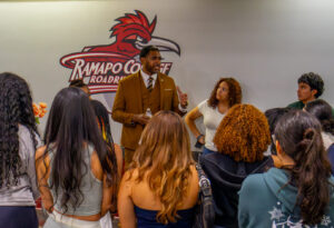 A man in a tan suit stands in front of and speaks to a group of students.